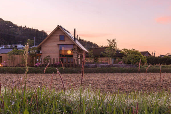 夕焼け空の田園風景の中に建つ、窓から見える照明と薪ストーブの煙突が印象的な三角屋根の木の家
